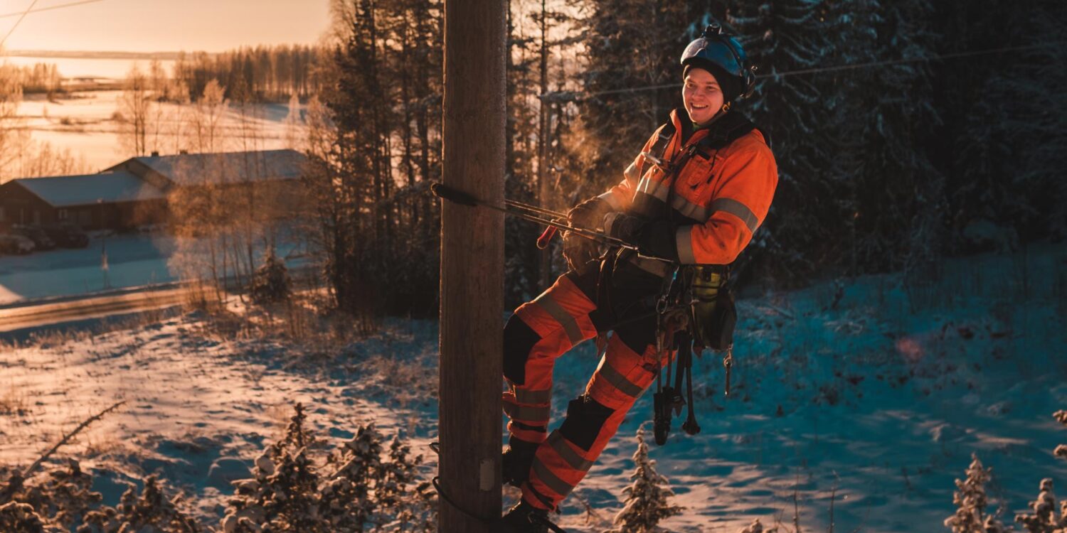 Verkostoasentaja sähköpylväässä. Taustalla talvimaisema ja ilta-aurinko paistaa.